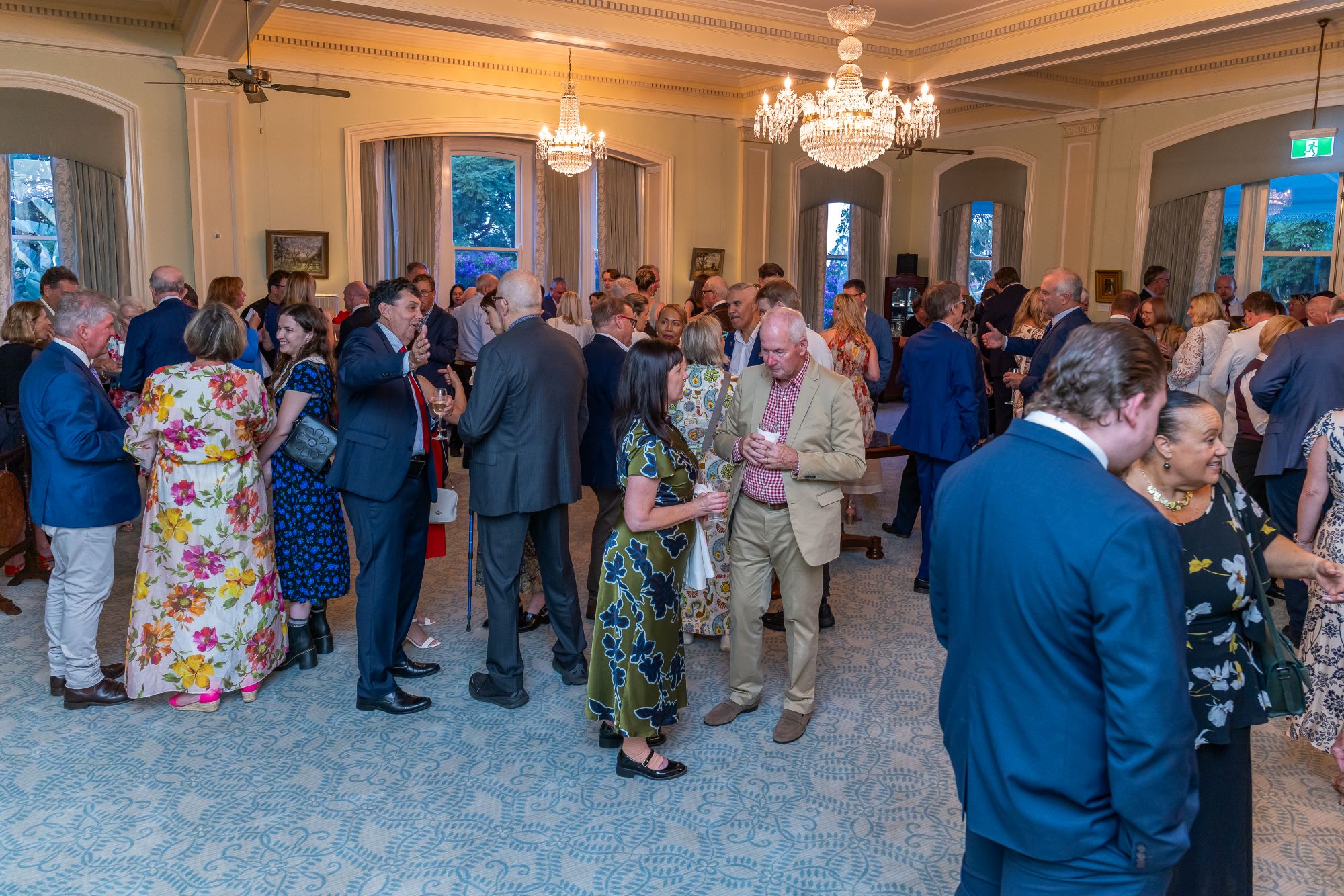 A room with a blue patterned carpet, on that carpet stands multiple people dressed for a cocktail event, they are all talking to each other. the room has two large chandeliers that are lit. They are all happy chatting and networking together