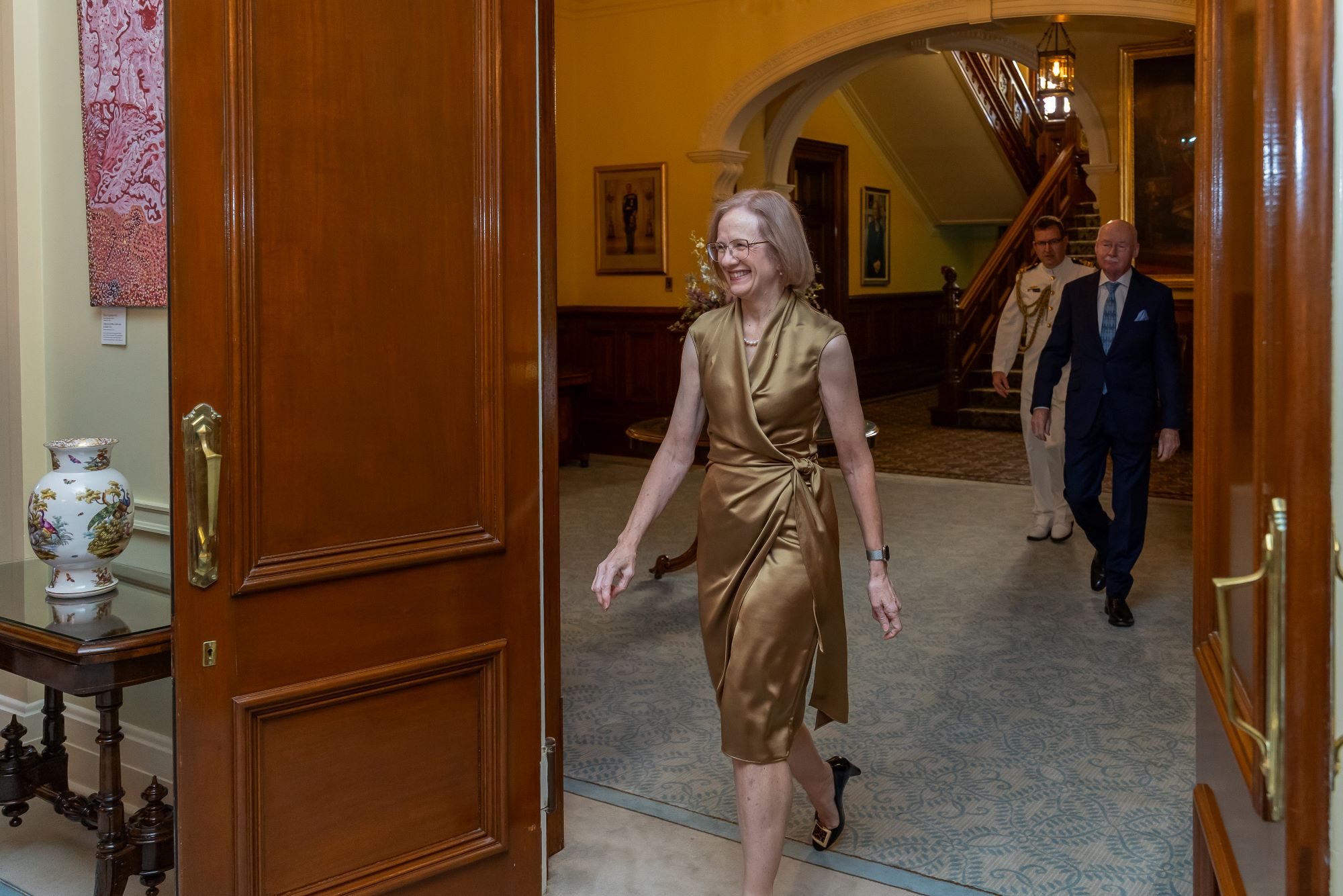 A lady in a gold dress is smiling and walking into another room, behind her is a man in a dark suit also walking in the same direction. They are both happy and smiling